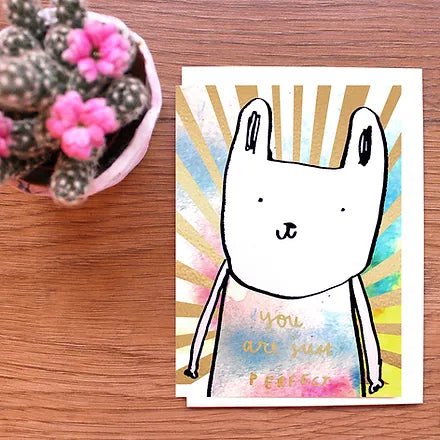 A greeting card with a hand-drawn illustration of a rabbit on a colorful background, placed on a wooden surface next to a small potted plant.