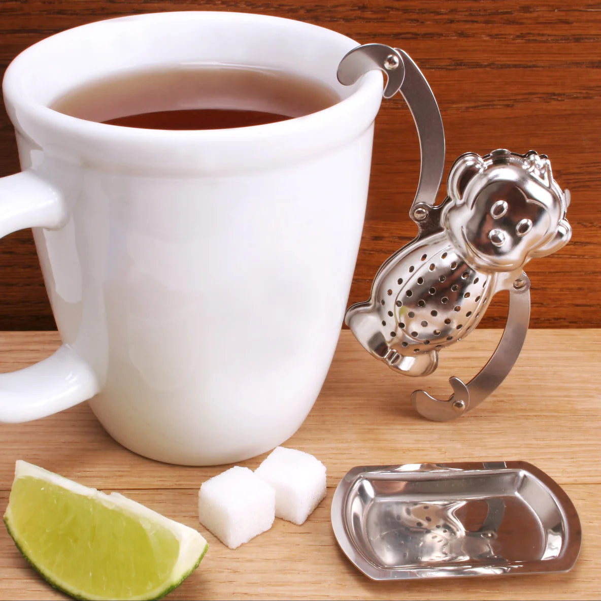 A stainless steel monkey shaped tea infuser hanging onto the side of a mug of tea with one of its arms. In front of the mug there is a little tray for the monkey to rest on and some sugar and lime.