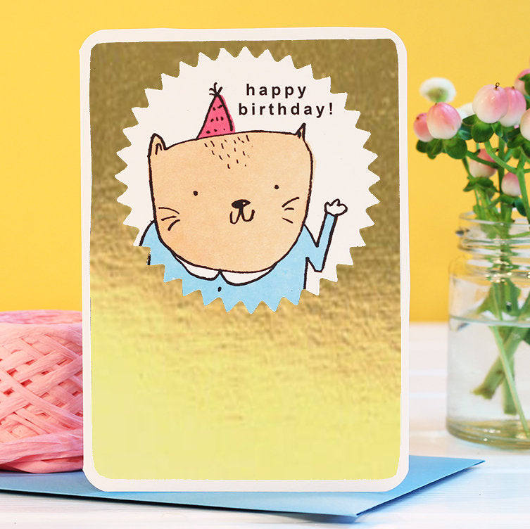 A greeting card featuring a hand-drawn illustration of a cat with a party hat, surrounded by a gold foil background. The text on the card reads 'happy birthday!'.