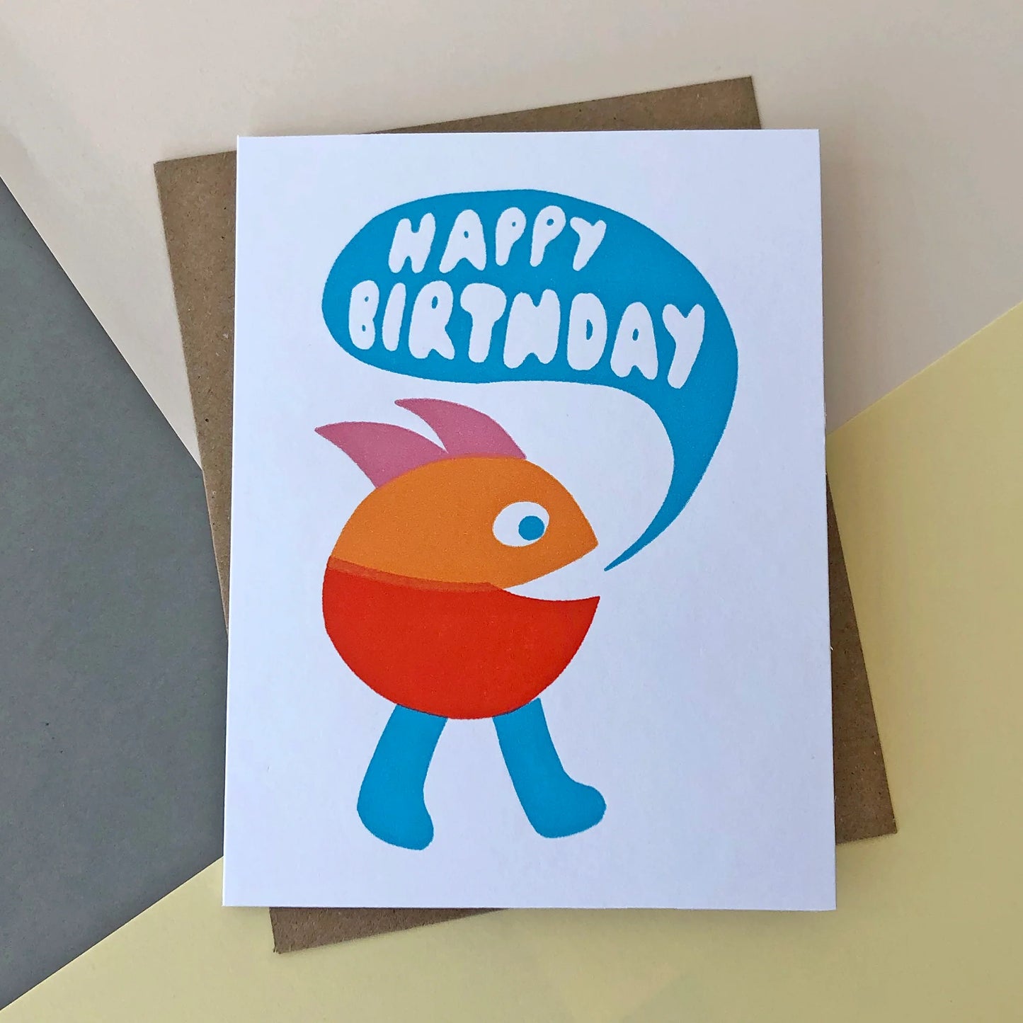 A white birthday card with a blue, orange and red pac-man like figure on the front with a pink mohican and legs. There is a speach bubble coming from him that reads 'Happy Birthday'