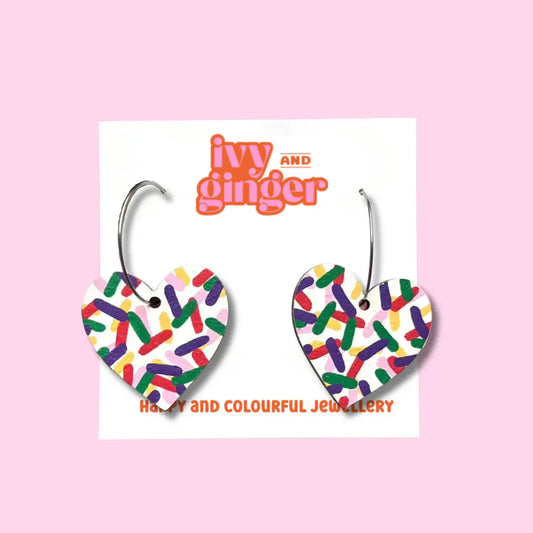 A pair of hand-painted wooden heart-shaped earrings with a multicoloured confetti design, displayed on a white backing card  with the brand name 'Ivy and Ginger' above.