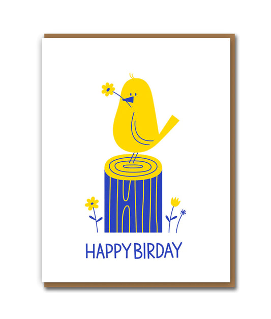 A white birthday card with a yellow bird stood on a blue felled tree. The bird has a yellow flower in its beak and there is text underneath that reads 'Happy Birday'