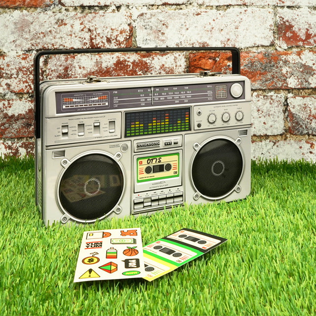 A metal lunchbox in the shape of a boom box. The lunchbox is in front of a wall with some stickers laid out in front of it on the grass.