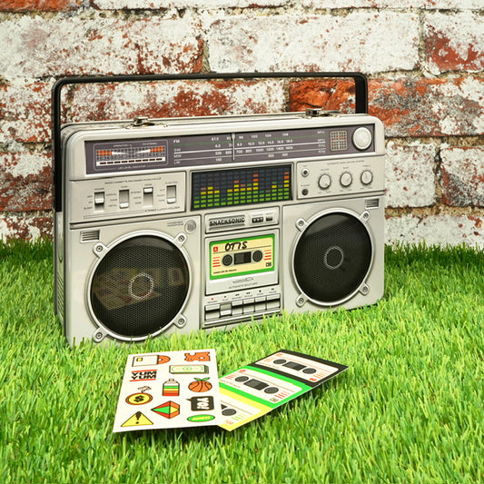 A metal lunchbox in the shape of a boom box. The lunchbox is in front of a wall with some stickers laid out in front of it on the grass. 