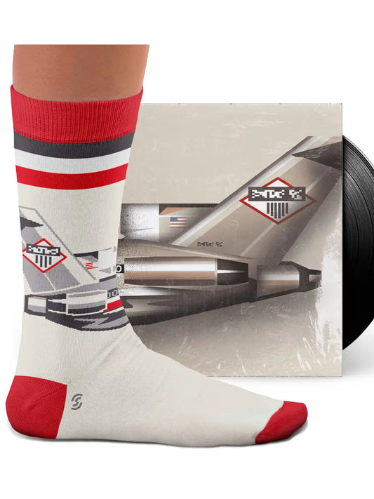 A person’s foot wearing a white sock with red and black stripes. On the front of the sock is a white black and red American Airlines Boeing 727 and it is resting on a vinyl record cover with the same logo.