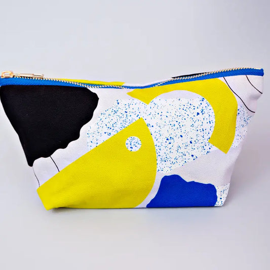 A cosmetic bag with black yellow and blue abstract shapes on. There is also a patch on the bag what is white with blue paint speckles all over. 