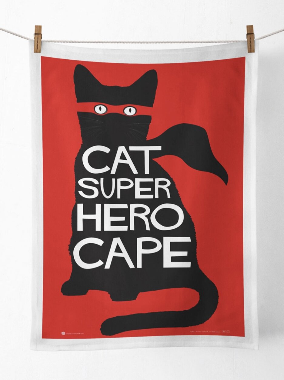 A red tea towel on a washing line. There is a black cat on the front wearing a red super hero mask and text on the tea towel reads: ' Cat super hero cape'