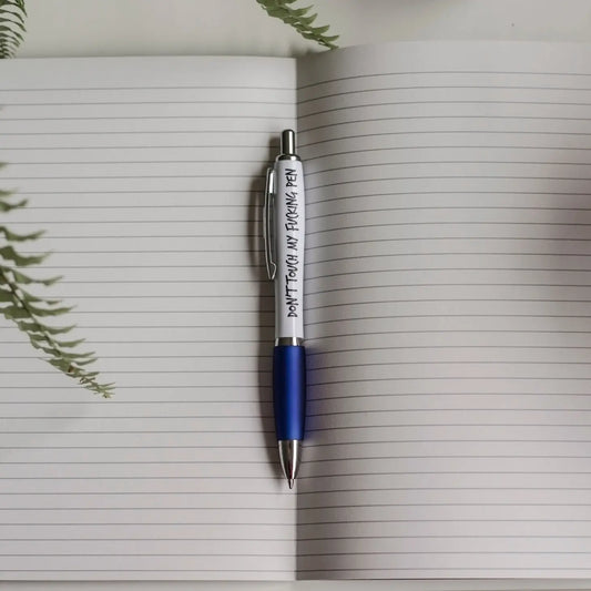 A white and blue pen laying in the middle of a notebook. The text on the pen reads: Don't touch my fucking pen 