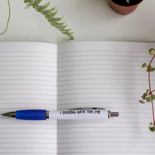 A white pen with blue accents lying on an open notebook. The pen has text on it, and there are plants in the background.