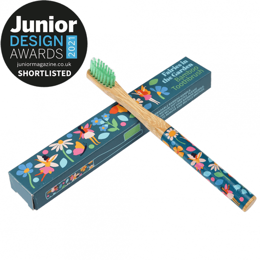 A wooden toothbrush with fairy images on the handle is leaning up against a blue cardboard box that reads: Faries in the garden bamboo thoothbrush. Junior Design awards 2021 shortlisted