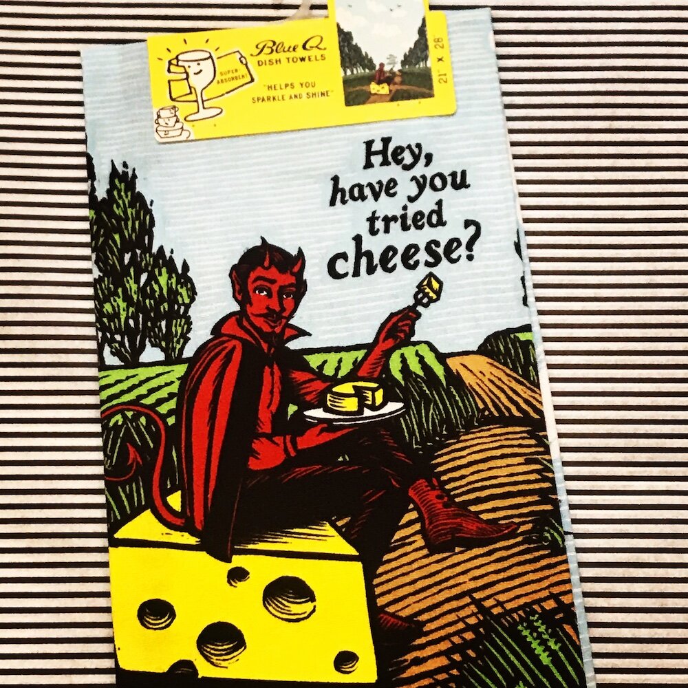 A Blue Q tea towel with the devil sat on a block of cheese in a field. 'Hey, have you tried cheese?'