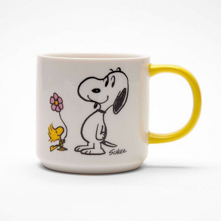 A white mug with a yellow handle. On the mug there is a graphic of Snoopy and Woodstock holding a flower.