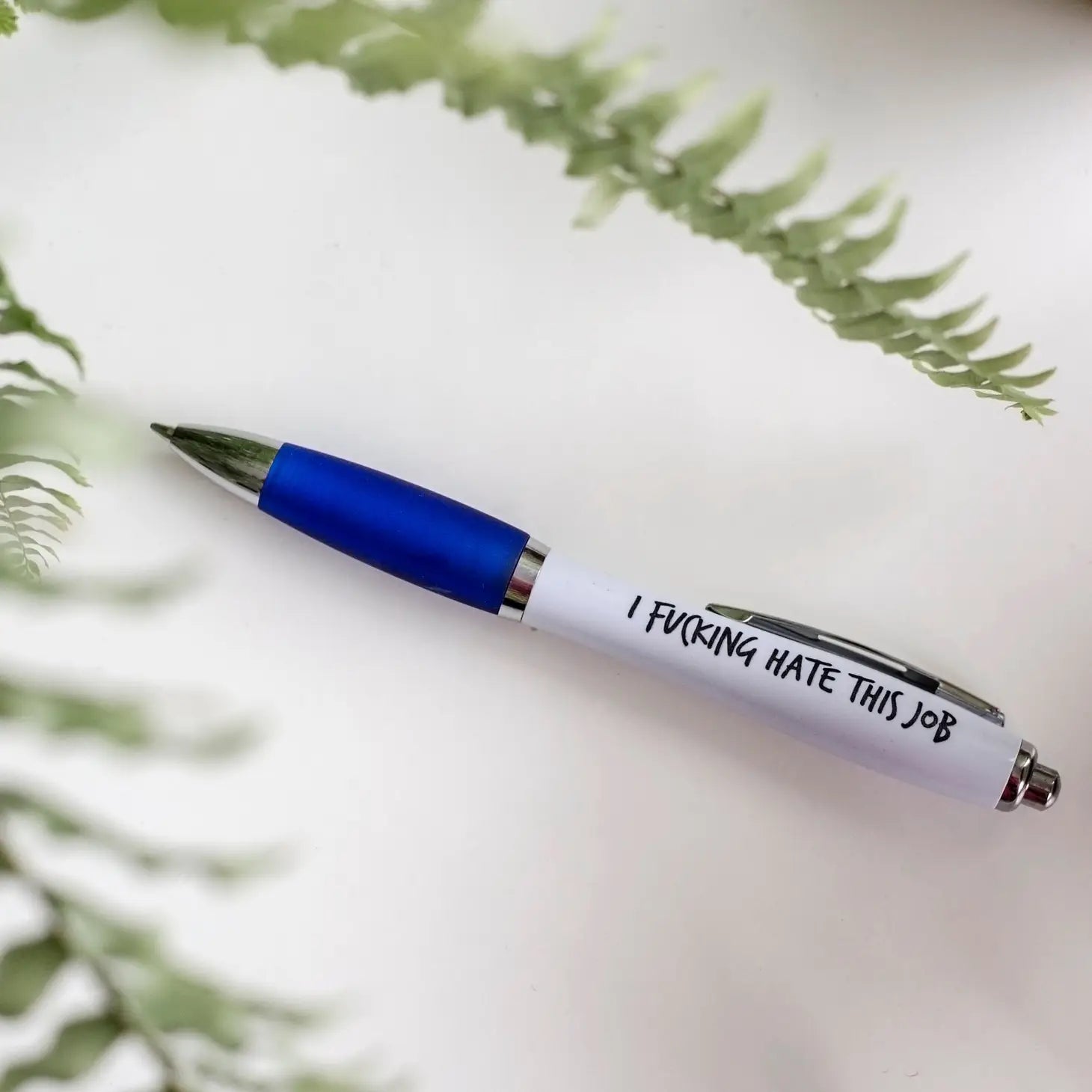 A white and blue pen laying on a table with foliage around. The text on the pen reads: I fucking hate this job