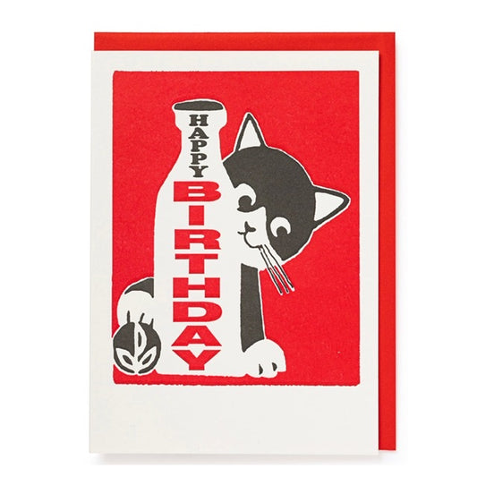 A white and red card with a bold block printed design on the front of a black and white kitten peering from behind a milk bottle that reads ' Happy Birthday' on the bottle