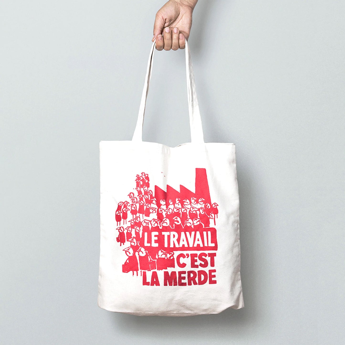 A white tote bag with a red drawings on of lots of grumpy people. The text on the bag reads: Le travail c'est la merde