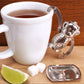 A stainless steel monkey shaped tea infuser hanging onto the side of a mug of tea with one of its arms. In front of the mug there is a little tray for the monkey to rest on and some sugar and lime. 