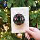 Hand holding a decorative Christmas ornament with 'Merry Christmas' text against a festive tree background.