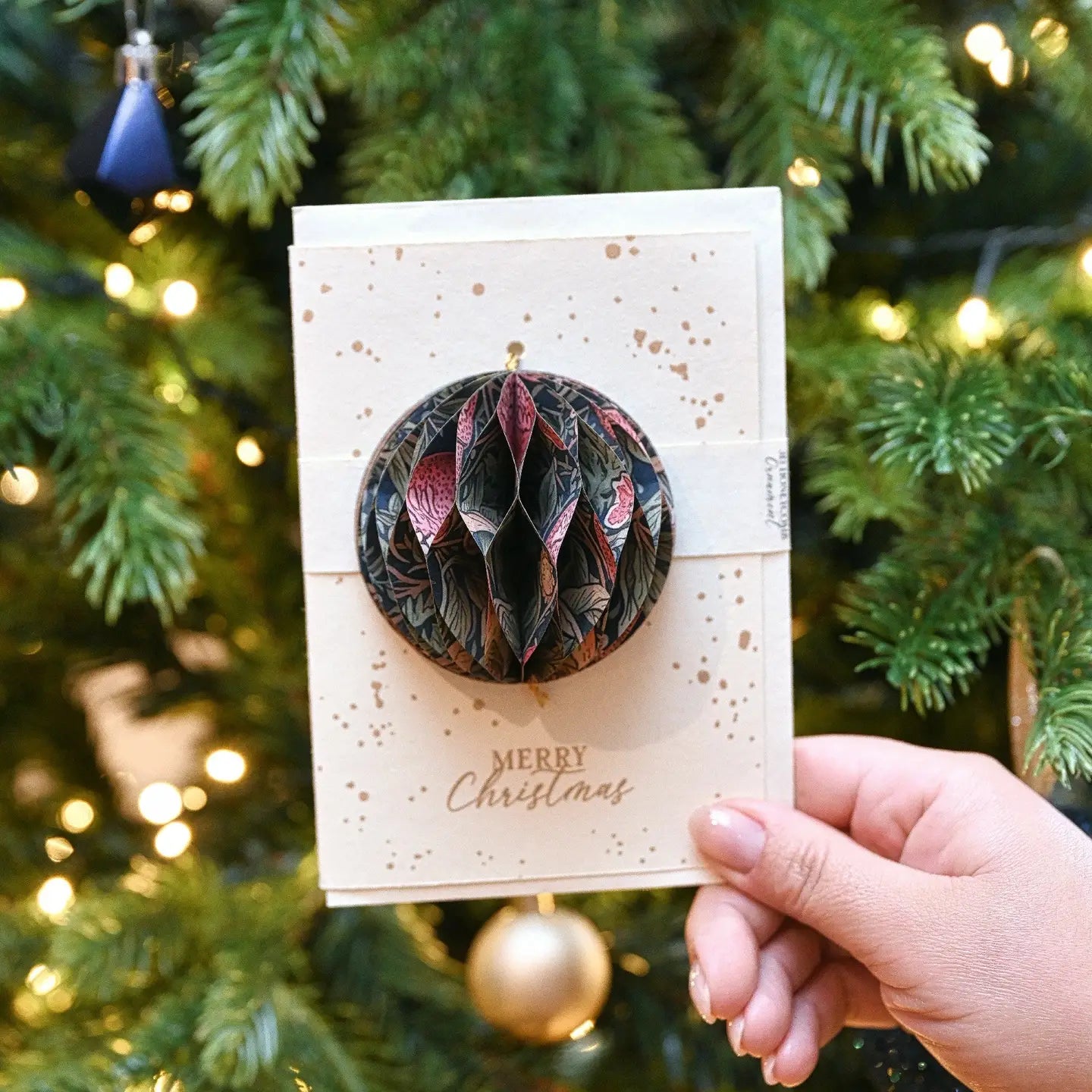 Hand holding a decorative Christmas ornament with 'Merry Christmas' text against a festive tree background.