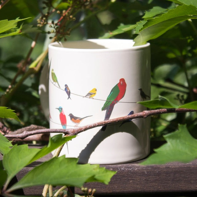 A cream mug with silhouettes of birds on a wire all over that is placed on some wood with tree branches all around 