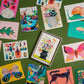 14 colourful postcards laid out on a table, they are different postcards containing pictures of animals or flowers. 