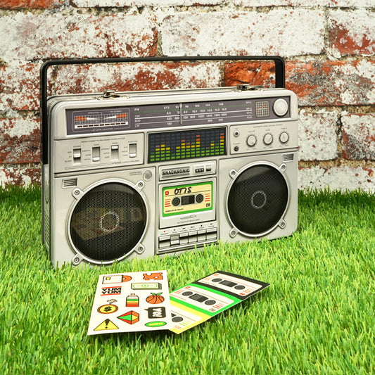 A metal lunchbox in the shape of a boom box. The lunchbox is in front of a wall with some stickers laid out in front of it on the grass.