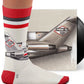 A person’s foot wearing a white sock with red and black stripes. On the front of the sock is a white black and red American Airlines Boeing 727 and it is resting on a vinyl record cover with the same logo.