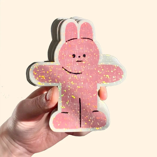 A hand holding a sticker in the shape of a pink rabbit with rainbow glitter.