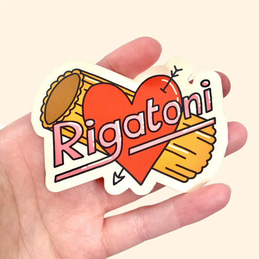 A hand holding a gloss vinyl sticker with a red heart and the word 'Rigatoni' written in pink and orange, resembling a tattoo style design.