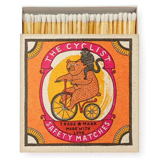An open box of matches. On the front of the matches there is a drawing of an orange bear riding a red bicicle with a black monkey on his shoulder. The box has an orange background with some fancy pink border. The text on the box reads: ' The Cyclist - Trade mark - Made with love - Safety matches ' 