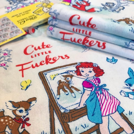 A Blue Q tea towel with a girl in an apron painting a picture of the deer with text that reads: 'Cute little fuckers'
