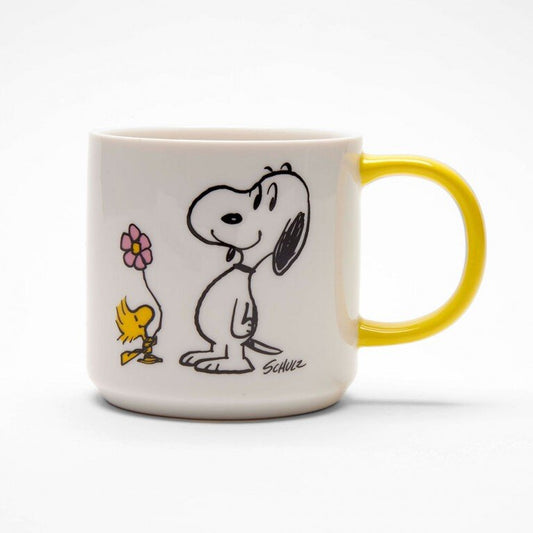 A white mug with a yellow handle. On the mug there is a graphic of Snoopy and Woodstock holding a flower. 