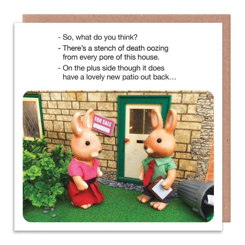 A white card with 2 rabbits stood outside of a house that is for sale. The text on the card reads: So, what do you think? - There's a stench of death oozing from every pore of this house. - On the plus side it does have a lovely new patio out back...