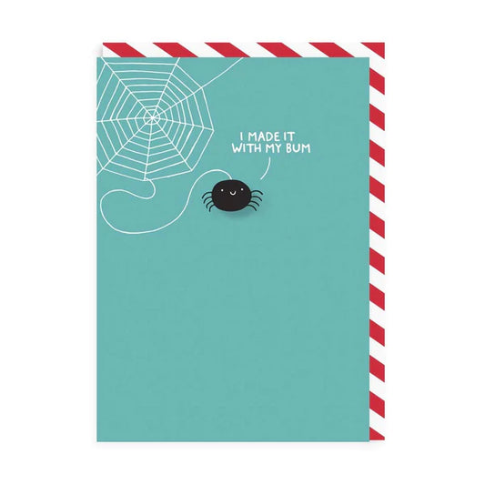 A blue card with a spider pin badge in the middle and a spiders web in the top corner. There is text on the  card coming from the spider that reads: I made it with my bum 